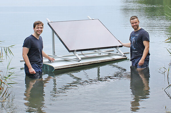 Zwei Männer stehen im Wasser eines Sees und halten stolz eine schwimmende Plattform mit einem Solarpanel.